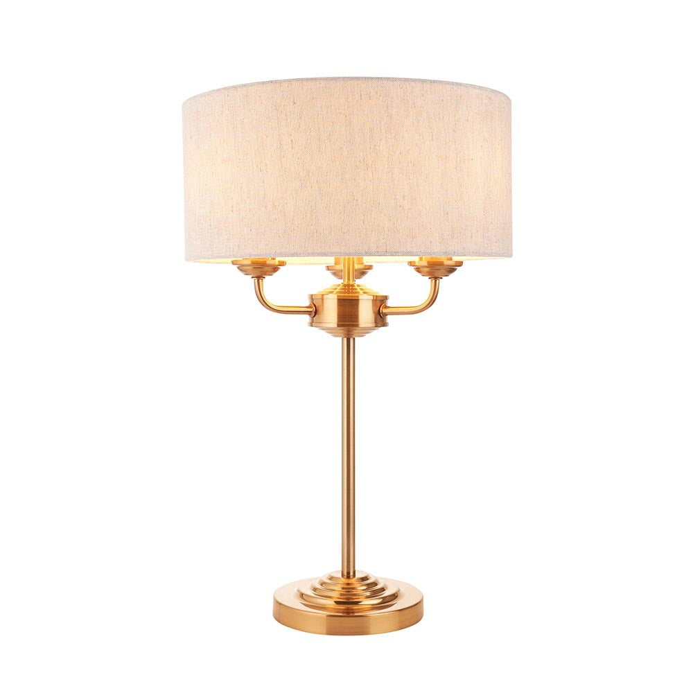 Brenley 3 Light Table Lamp in brass, with a stepped round base, tall stem, three arms, and a drum-shaped linen shade on a white background.