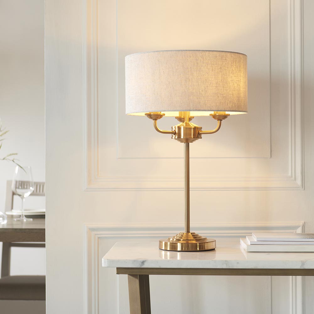 Brenley 3 Light Table Lamp in brass with a stepped base and column, topped by a grey linen drum shade, styled on a marble console table.