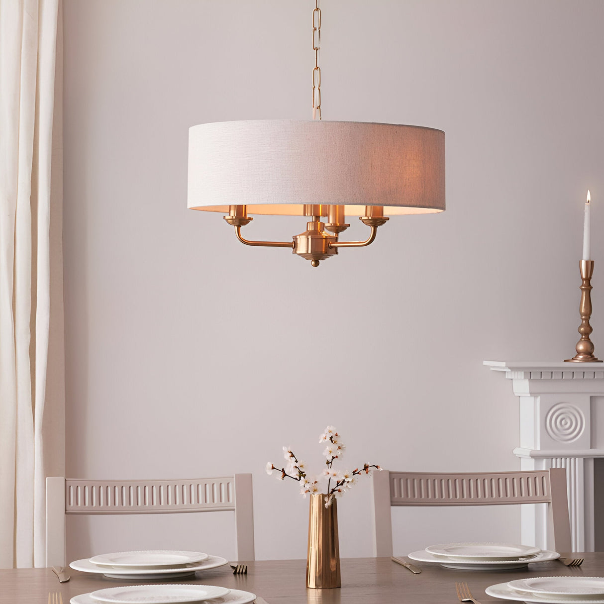 Brenley 3 Light Pendant in vintage brass with a natural linen drum shade, hung above a dining table set with white plates and a brass vase.