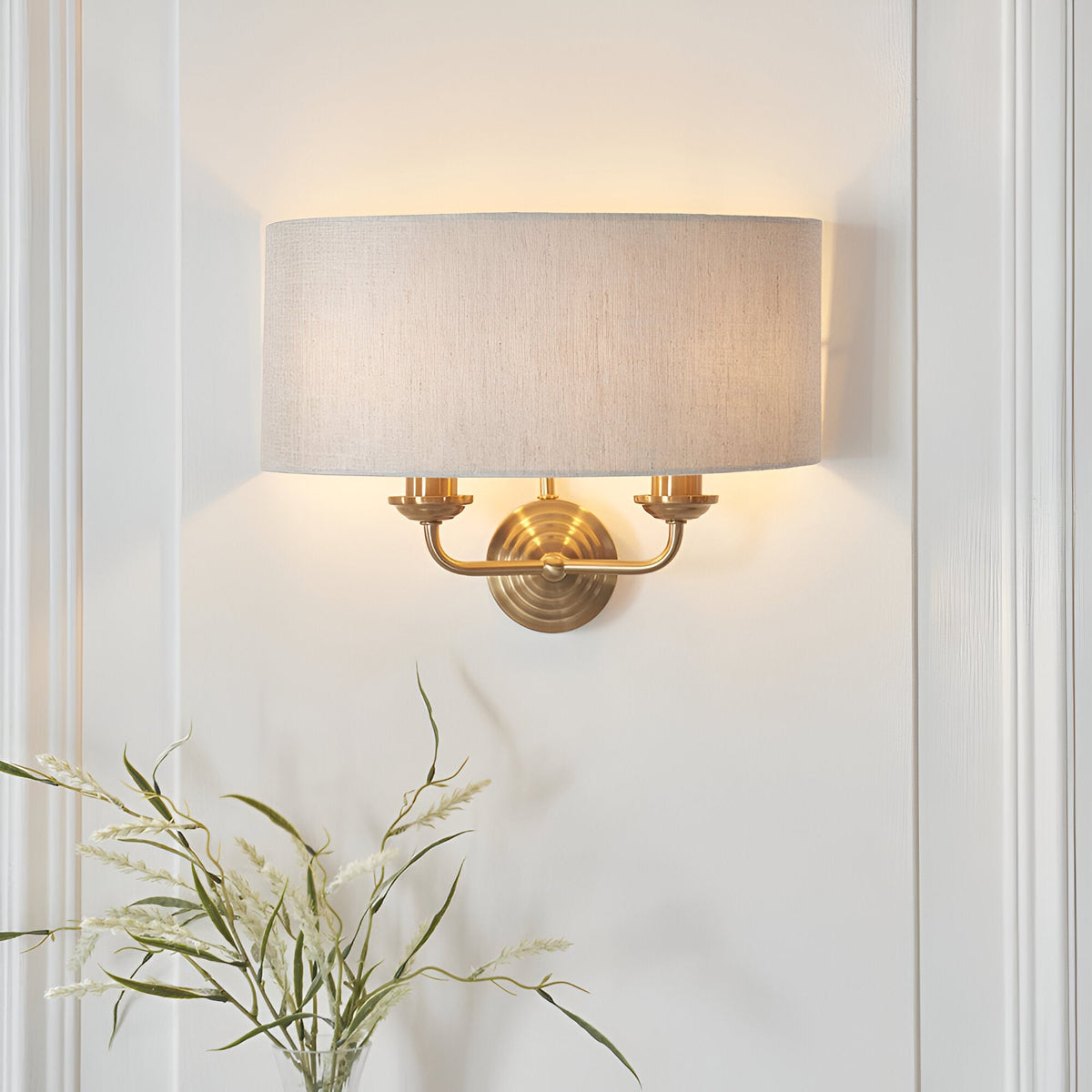 Endon Brenley 2 light wall light in vintage brass with a natural linen drum shade, mounted on a white panelled wall above a vase of dried grasses.