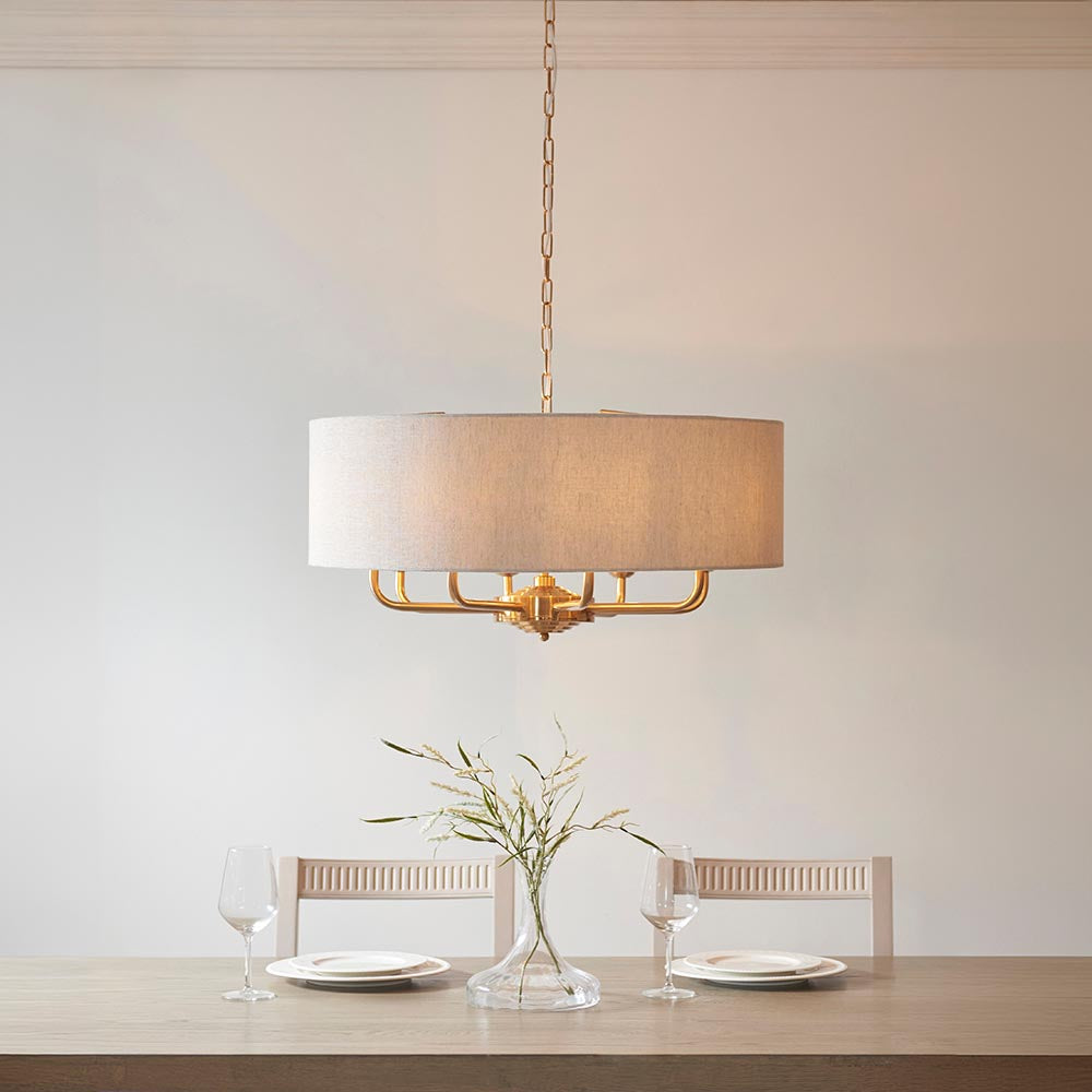 Endon Brenley pendant with wide natural linen drum shade on vintage brass frame, hanging above a dining table set with wine glasses and a vase.