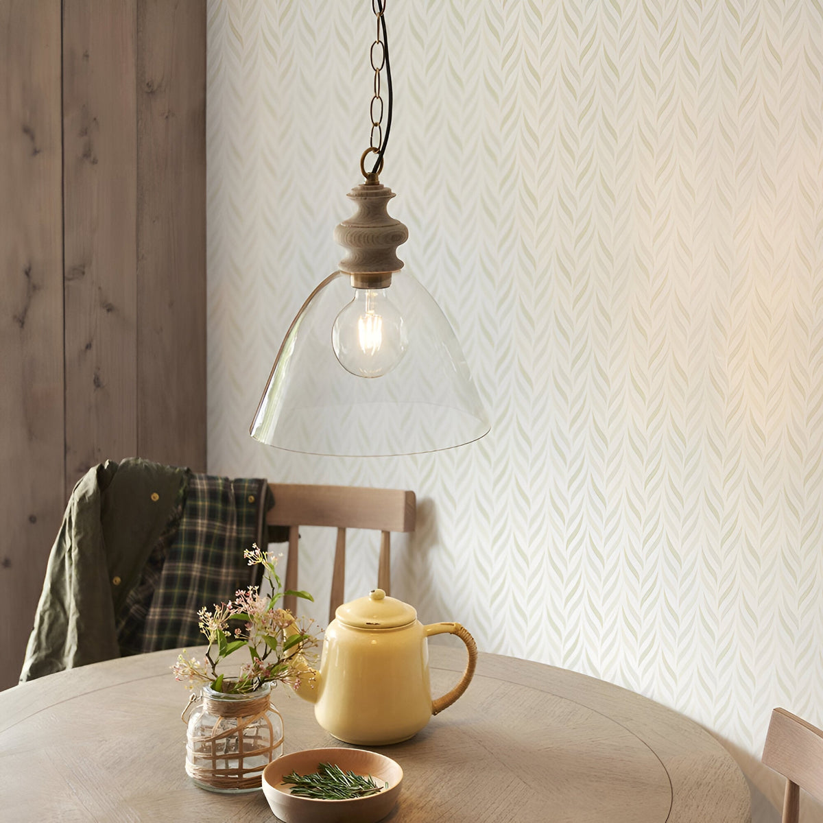 Bevwick pendant light with clear flared glass shade and natural ash wood top on brass chain, hung above a dining table in a kitchen setting.