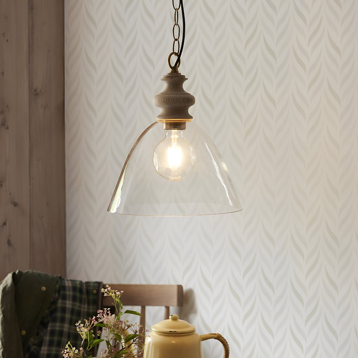 Bevwick pendant light with flared clear glass shade, turned ash wood top and black chain, shown lit above a dining table with wallpaper backdrop.