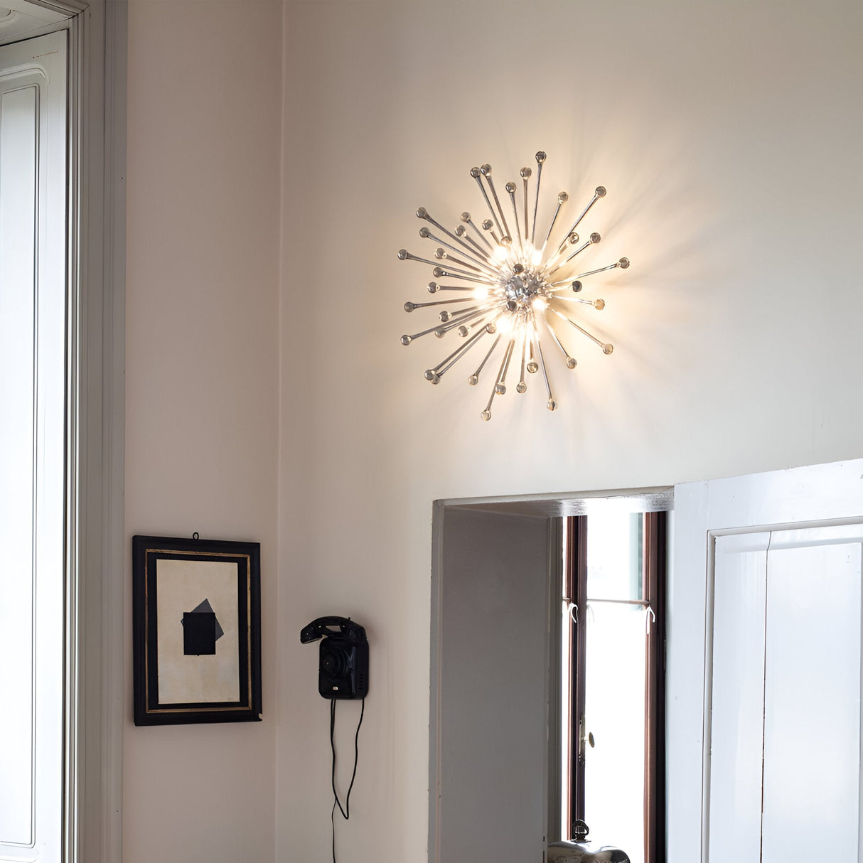 A contemporary Wander 5 Light Ceiling & Wall Light in chrome, featuring transparent glass elements, brightens the room and casts a glow over the vintage telephone mounted below it. Nearby, a framed abstract artwork adorns the white walls, while a tall window allows partial sunlight to filter in.