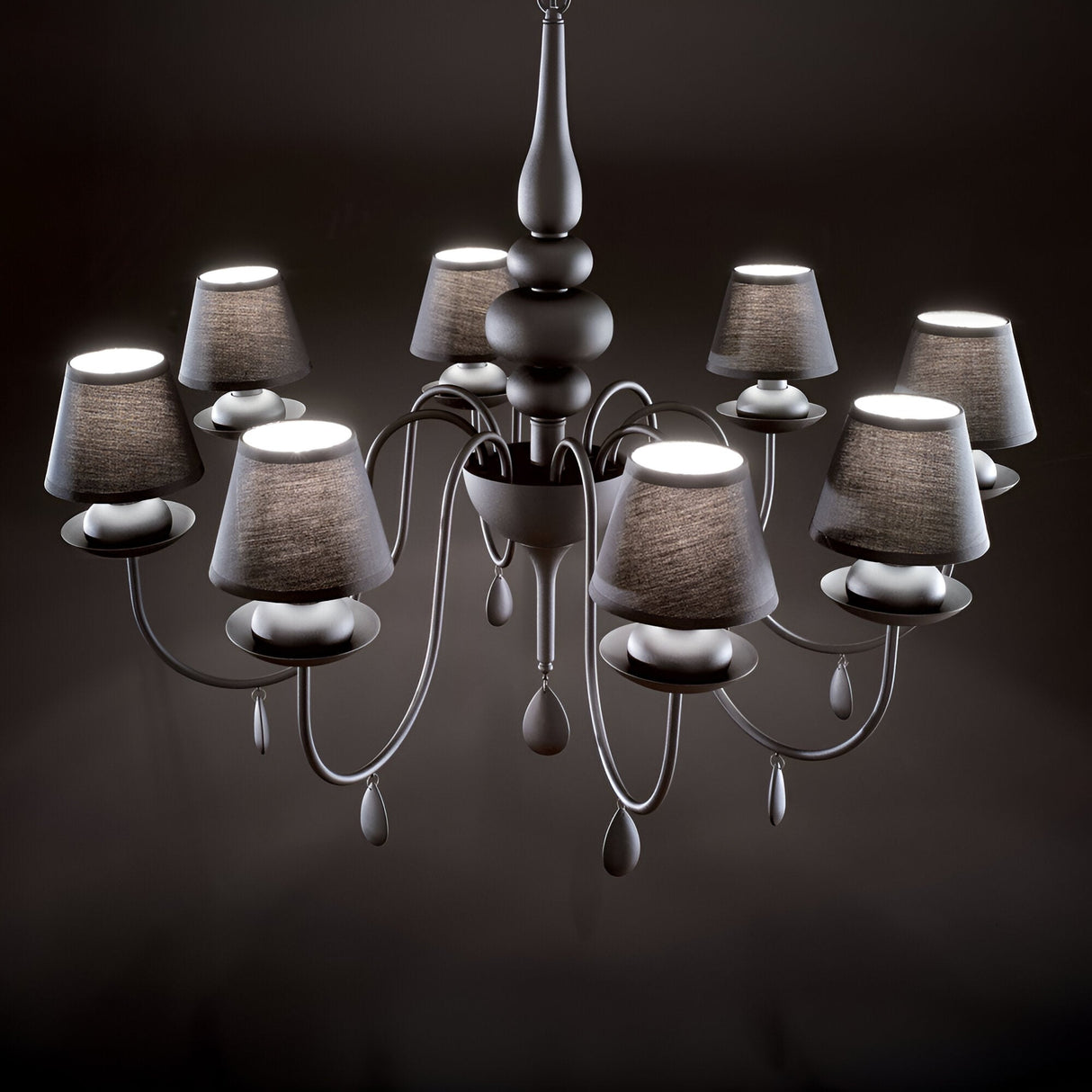 The Wisp 8 Light Chandelier - Black serves as a decorative centerpiece with its black satin enamel finish, featuring eight lamps adorned with black shades. Its ornate central structure and gracefully curved arms form an elegant silhouette against a dark background.