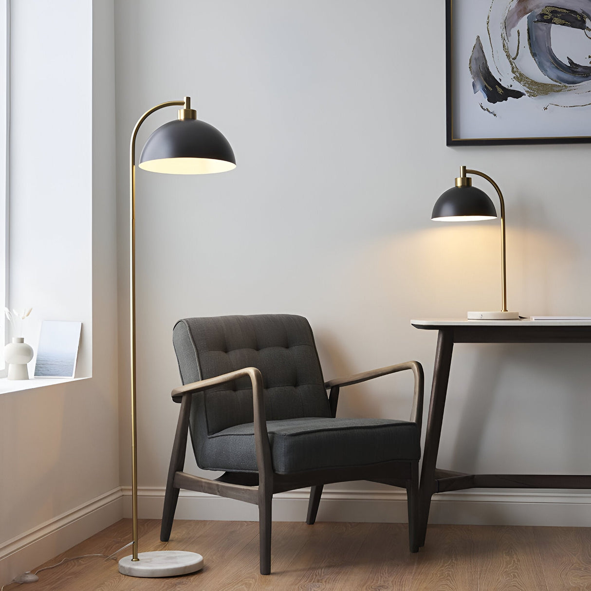Endon Ashmont table lamp with antique brass arched stem, matt black dome shade and marble base on a desk beside a grey armchair and floor lamp.