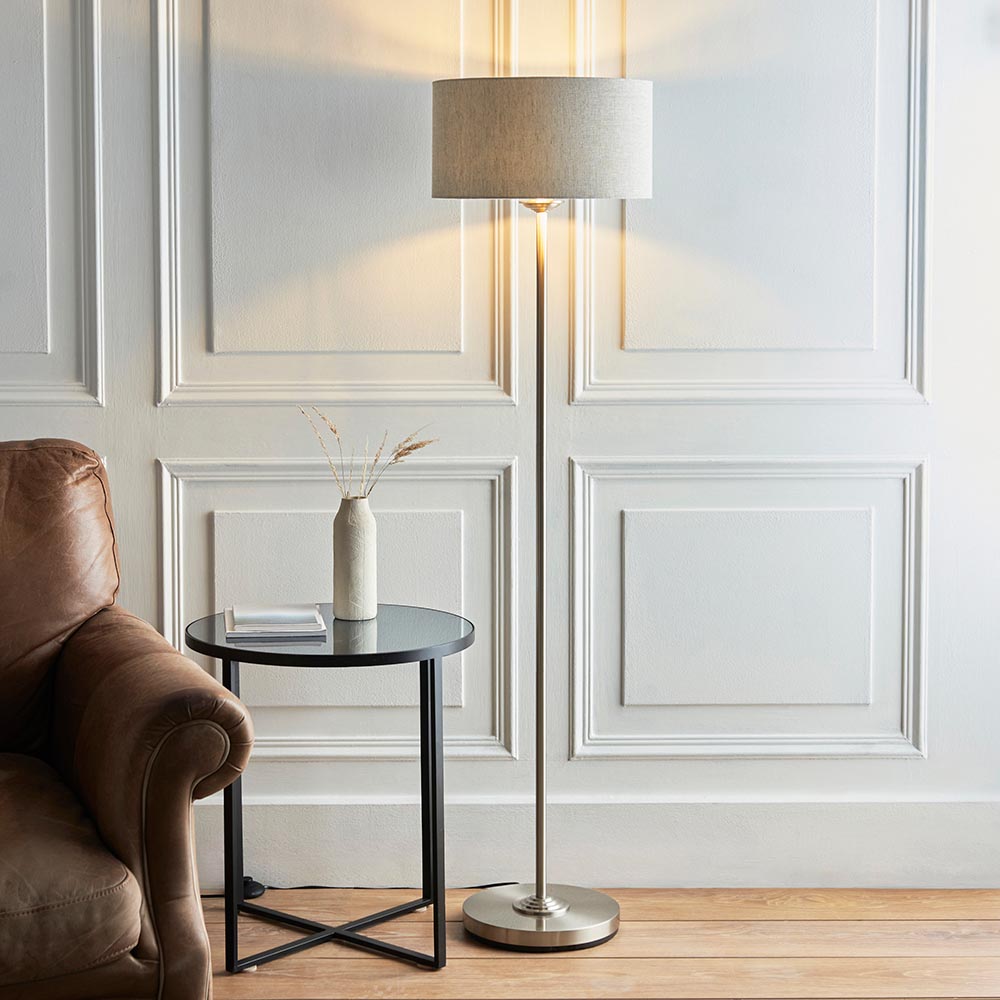 Endon Brenley floor lamp in chrome with a natural linen drum shade, lit beside a leather armchair and black side table in a panelled living room.