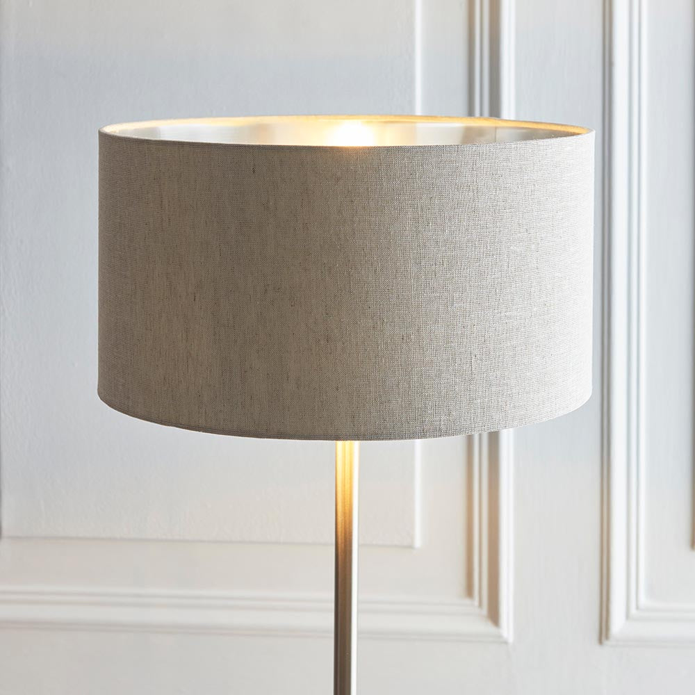 Endon Brenley floor lamp shown lit with a natural linen drum shade, metallic inner and slim chrome stem against a panelled off-white wall.