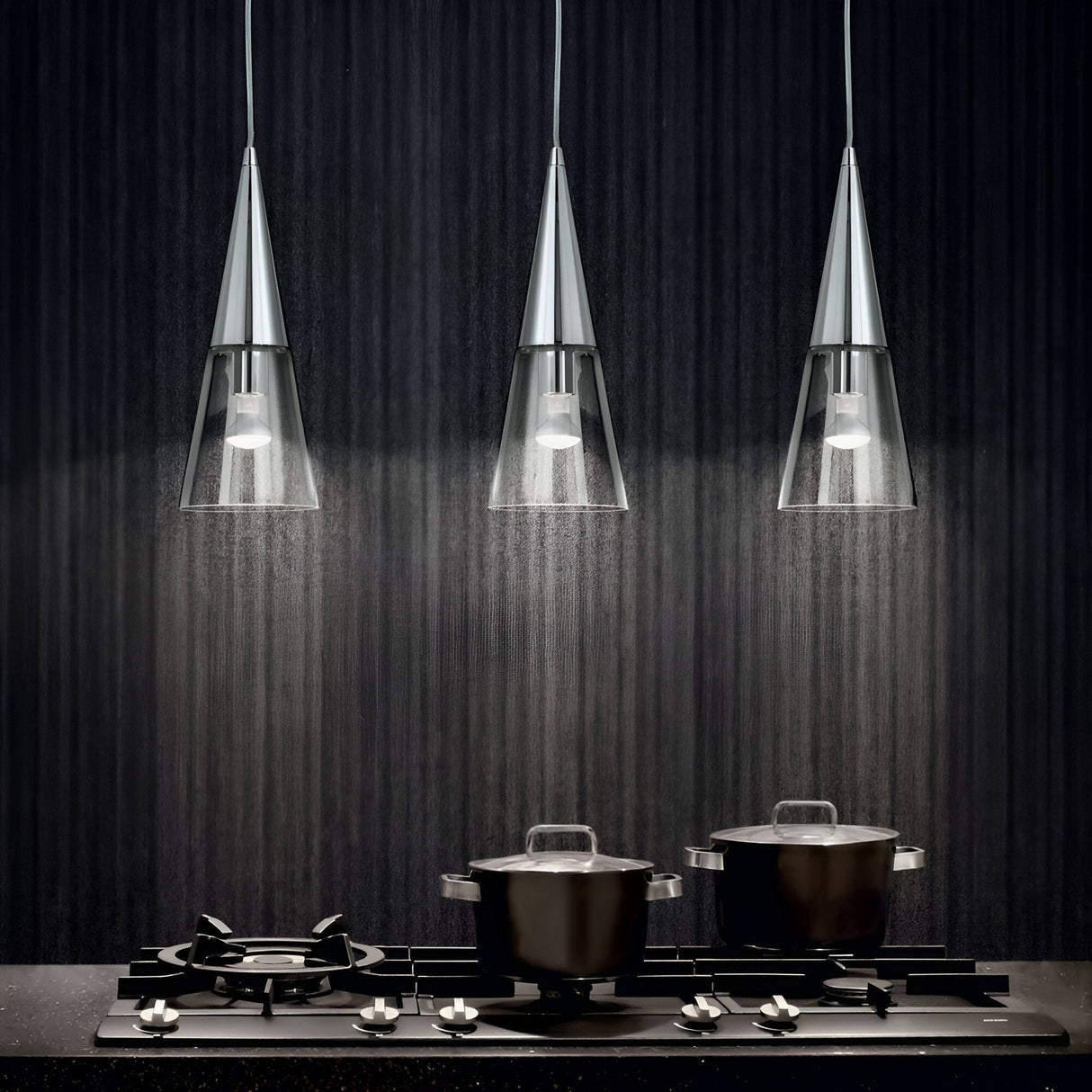 In a contemporary kitchen setting, a Cone 3 Light Bar Pendant - Silver with elegant clear glass cone-shaped shades provides illumination over a stovetop. Beneath it, two black pots with lids sit on different burners of a gas stove, framed by a dark textured wall. The dimmable bulbs enhance the versatility of the lighting ambiance.