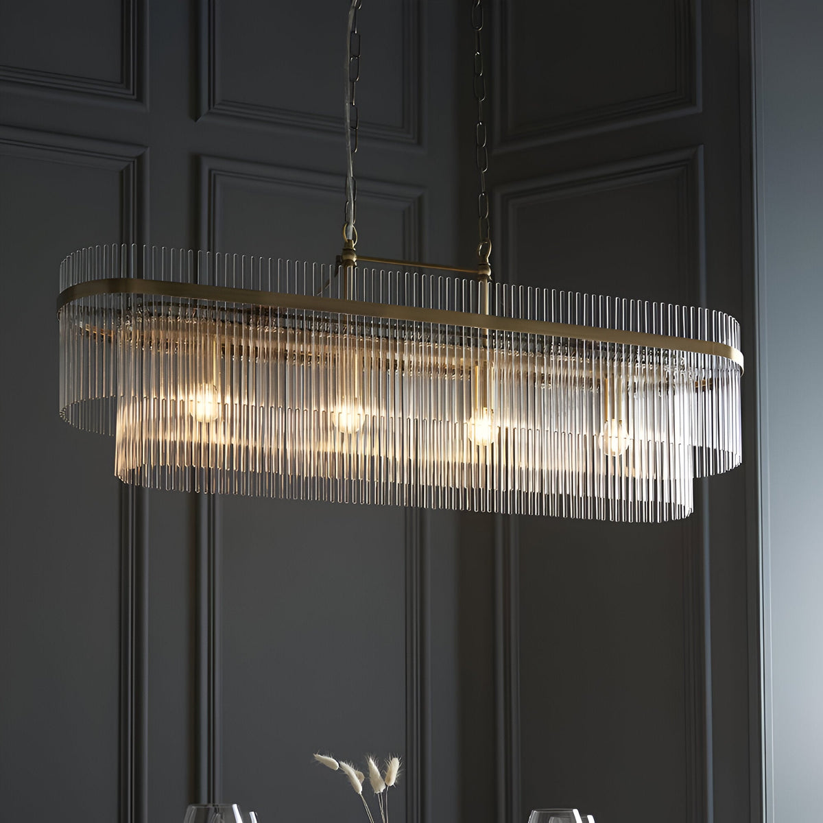 Beaumont 4 Light Linear Pendant in antique brass, an oval fixture lined with clear glass rods and four exposed bulbs, hung from chains in a dark panelled room.