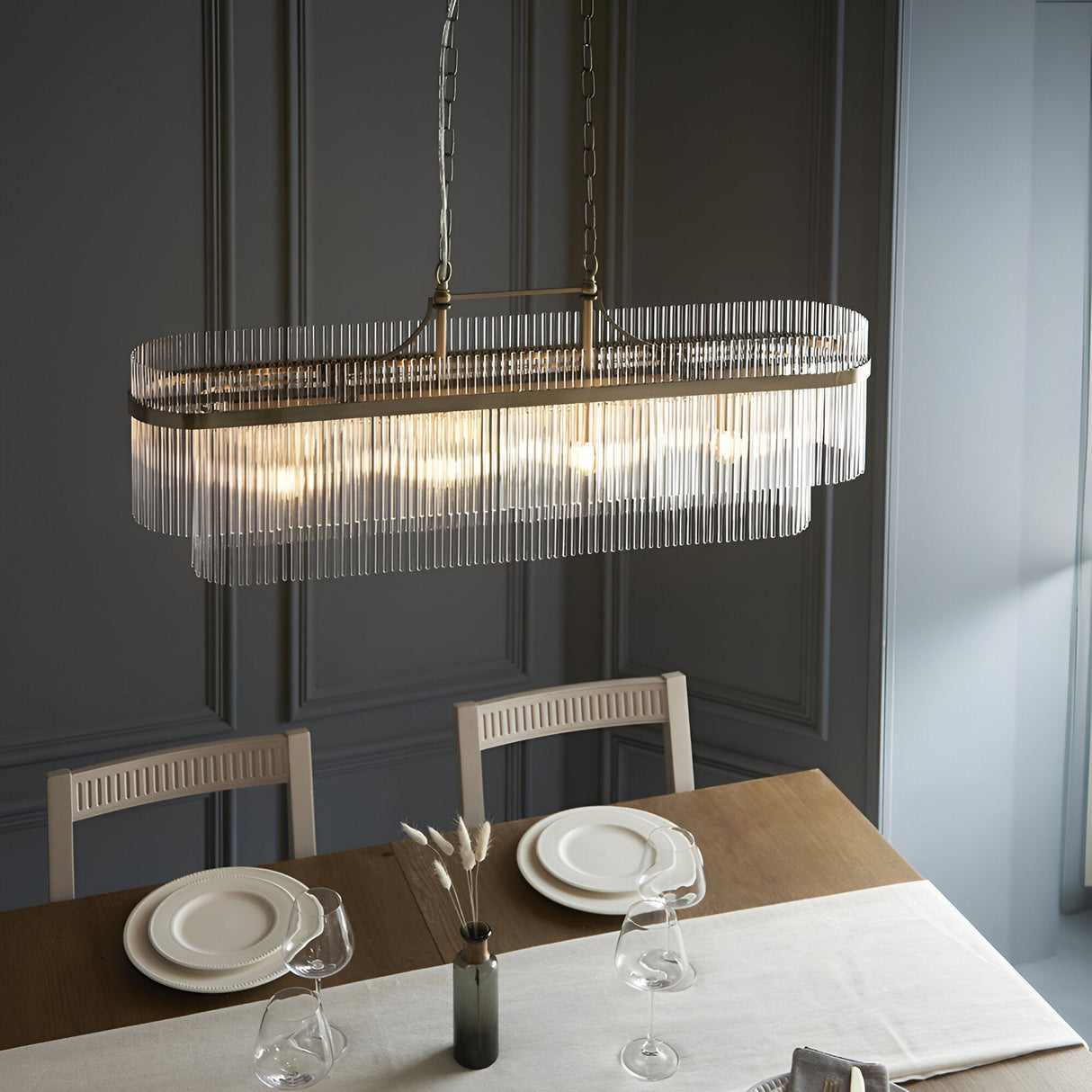 Beaumont 4 Light Linear Pendant in antique brass with clear glass rod fringe, hung above a dining table in a dark panelled room.