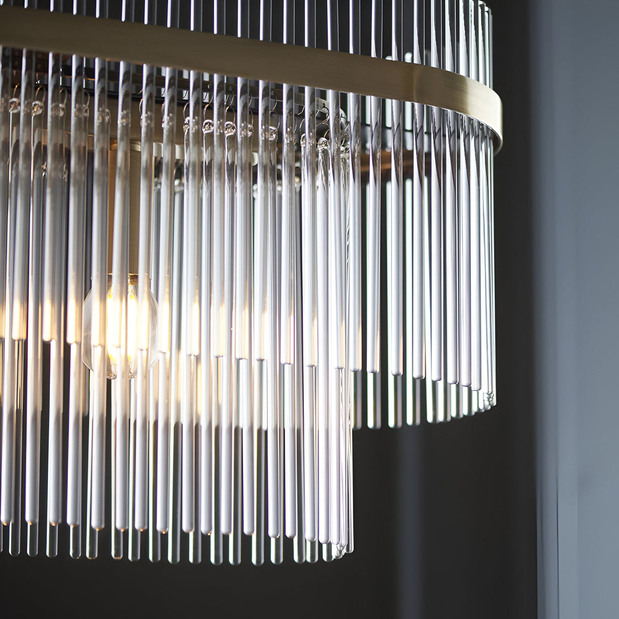Close-up of the Beaumont 4 Light Linear Pendant in antique brass, showing clear glass rods hanging from the brass bar with a visible bulb glow.