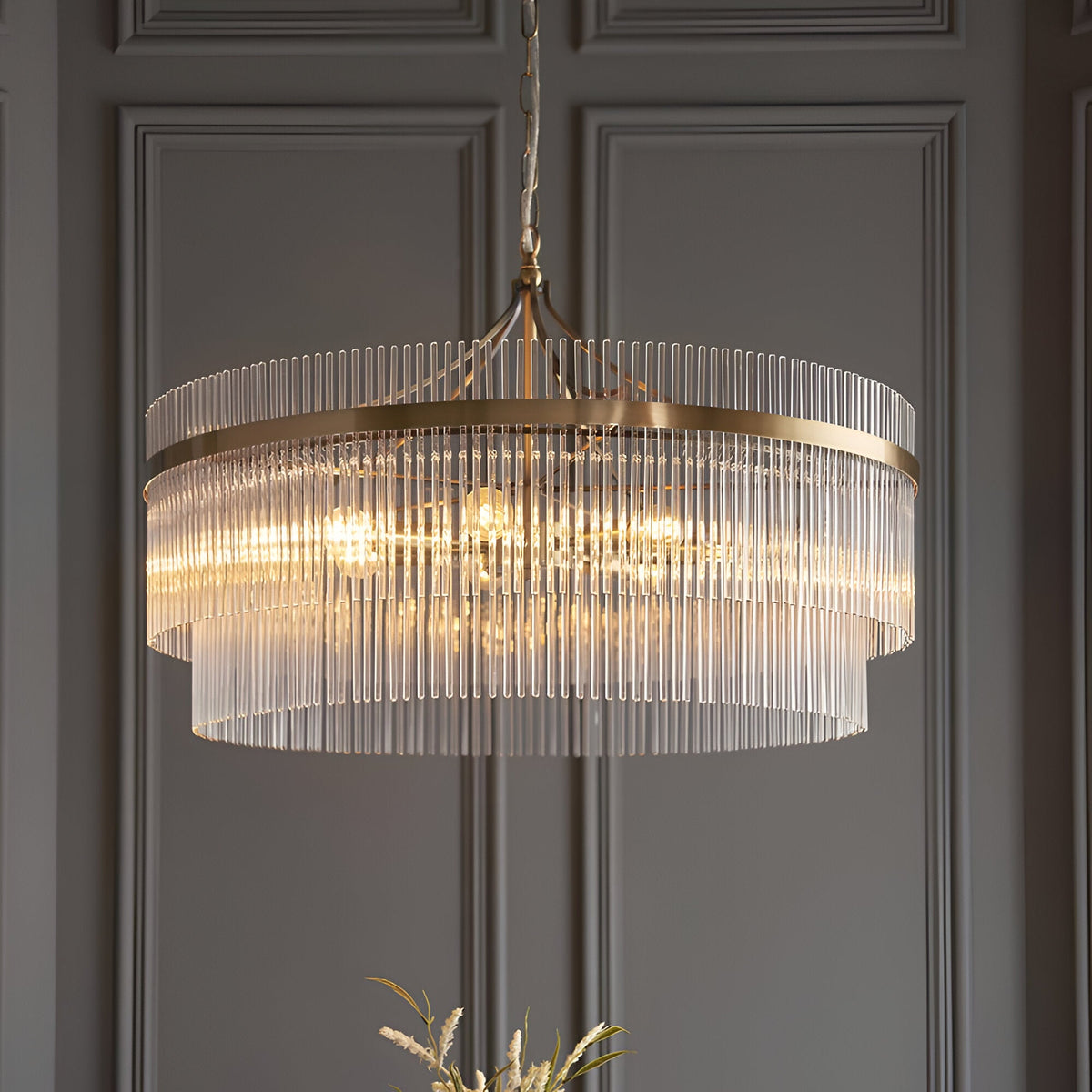 Endon Beaumont 7 Light Pendant in antique brass with tiered clear glass rods, hung from a chain against a panelled grey wall.