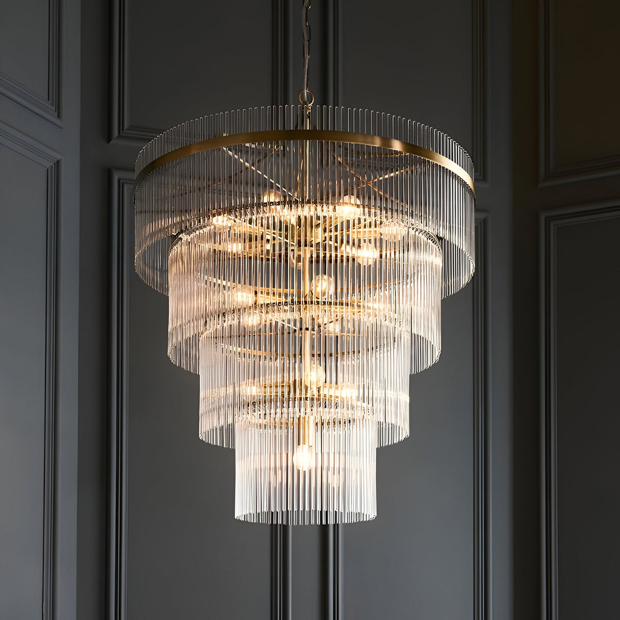 Beaumont 16 Light Chandelier in antique brass, a large tiered fitting with clear glass rods and exposed bulbs, hung against a dark panelled wall.