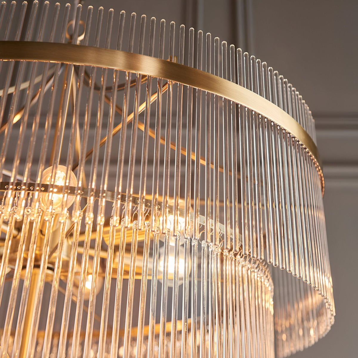 Close-up of the Beaumont 16 Light Chandelier in antique brass, showing tiered clear glass rods and exposed bulbs around a curved brass band.