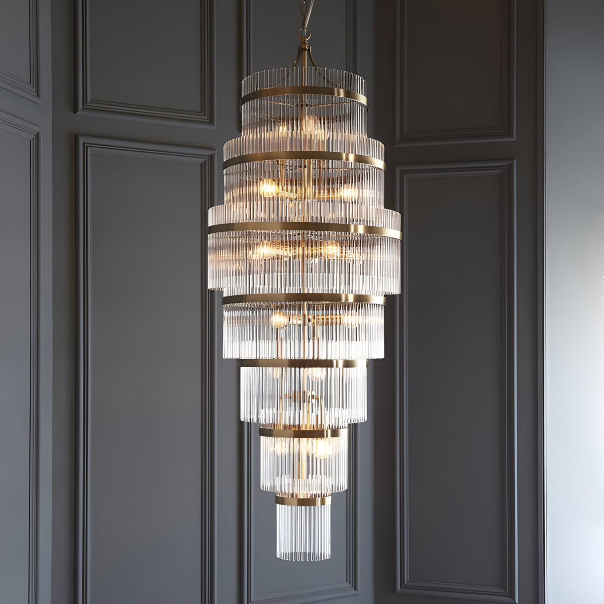 Endon Beaumont 21 light chandelier in antique brass with multi-tiered clear glass rods, hung from a chain against a grey panelled wall.