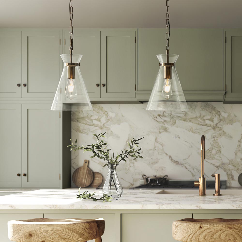 Endon Brenbury pendant lights in brushed brass with clear glass conical shades and exposed bulbs, hung as a pair above a marble kitchen island.