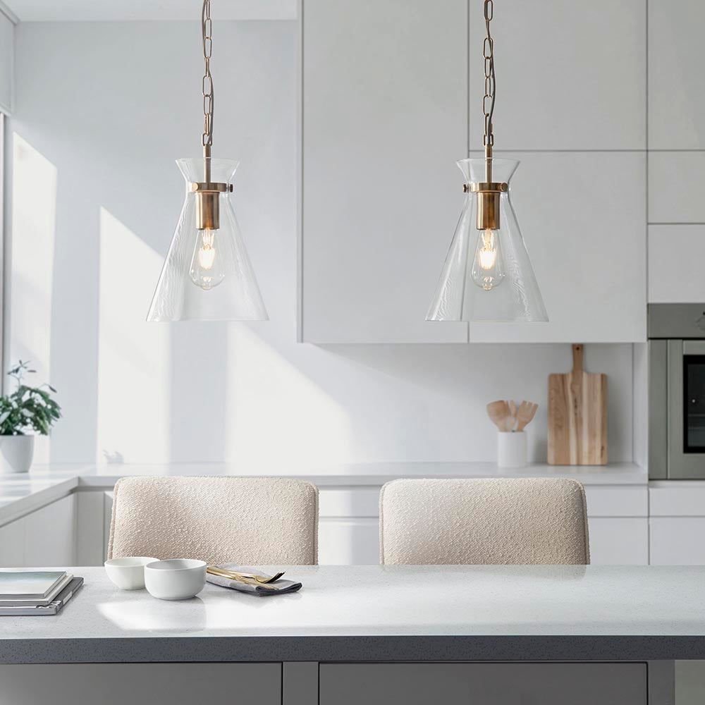 Two Endon Brenbury pendant lights in brushed brass with clear tapered glass shades, suspended by chains above a white kitchen island with boucle stools.