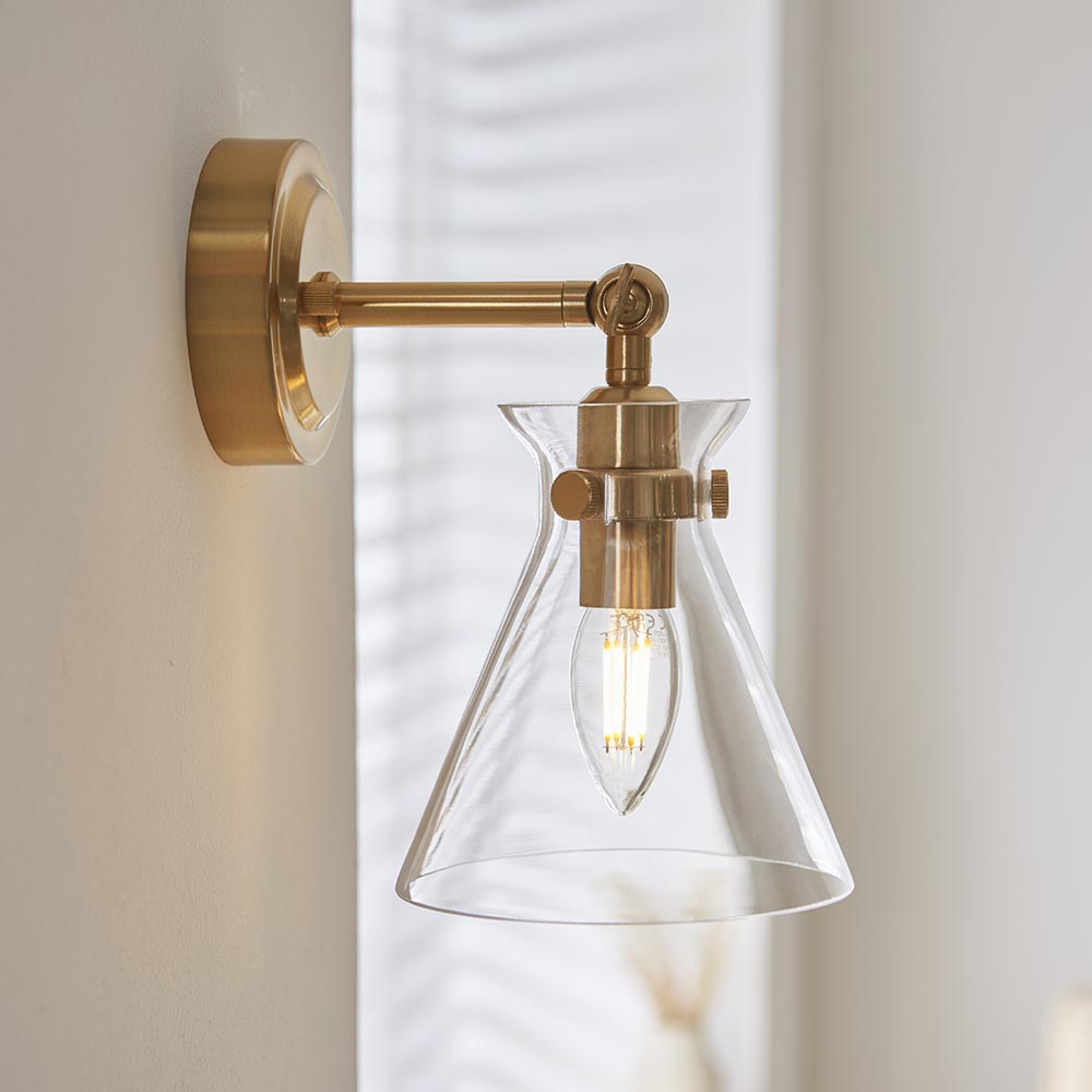 Endon Brenbury wall light in antique brushed brass with adjustable knuckle, clear glass conical shade and exposed E14 bulb, mounted beside a window.
