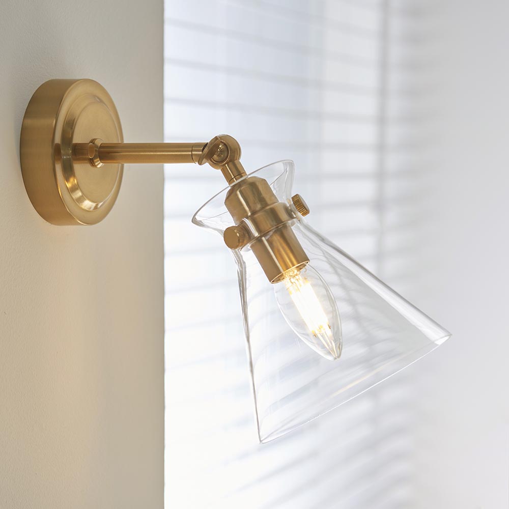 Endon Brenbury wall light in antique brushed brass with adjustable knuckle and clear glass cone shade, mounted beside a window with blinds.