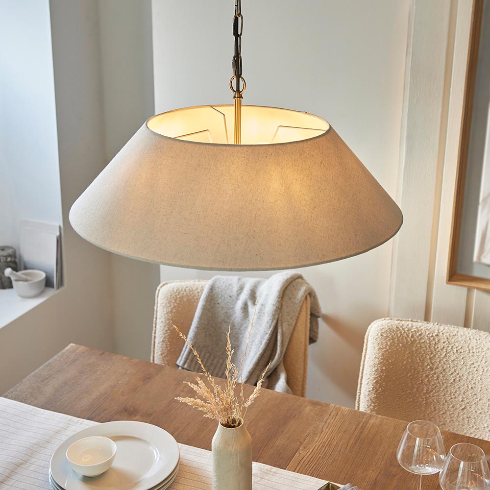Caldmere 5 Light Pendant in brass and natural linen, wide tapered shade hanging on brass chain above a wooden dining table in a light neutral room.