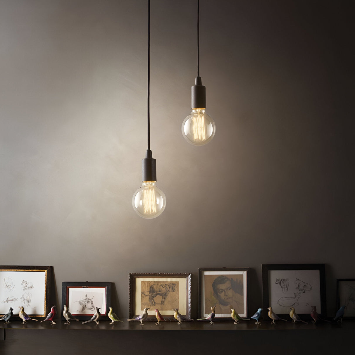 Two Ice 1 Light Pendants in black illuminate a shelf adorned with framed artwork and miniature bird figurines. The pendant lights' warm glow creates a cozy ambiance against the neutral wall, embodying a minimalist design that complements this contemporary style.