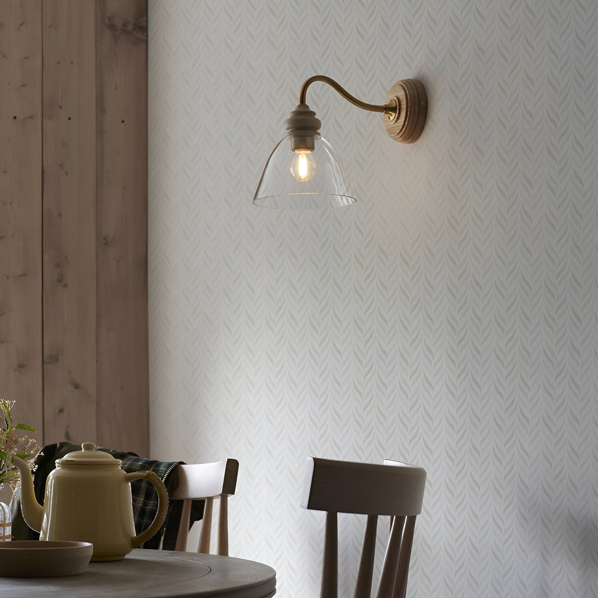 Endon Bevwick wall light in natural ash with aged brass arm and clear flared glass shade, mounted above a wooden dining table in a rustic interior.