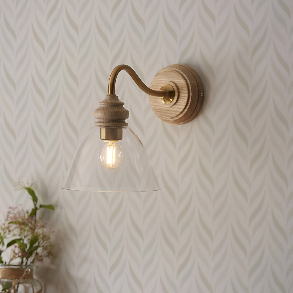 Endon Bevwick wall light in natural ash with aged brass arm and flared clear glass shade, mounted on patterned leaf wallpaper.