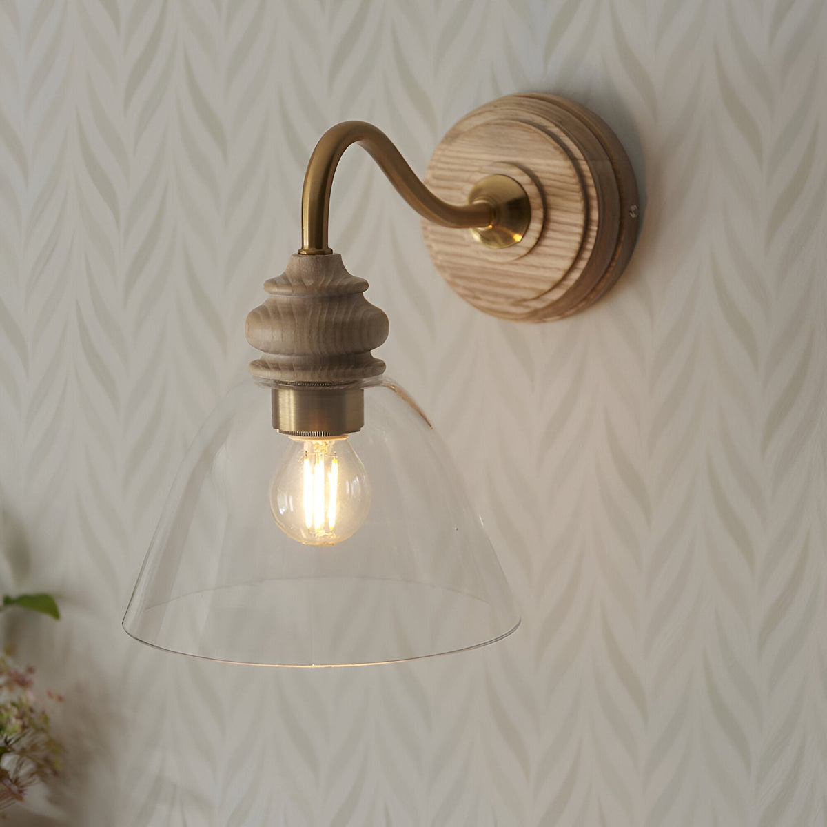 Endon Bevwick wall light in natural ash with aged brass arm and flared clear glass shade, lit E14 bulb, on leaf-patterned wallpaper.
