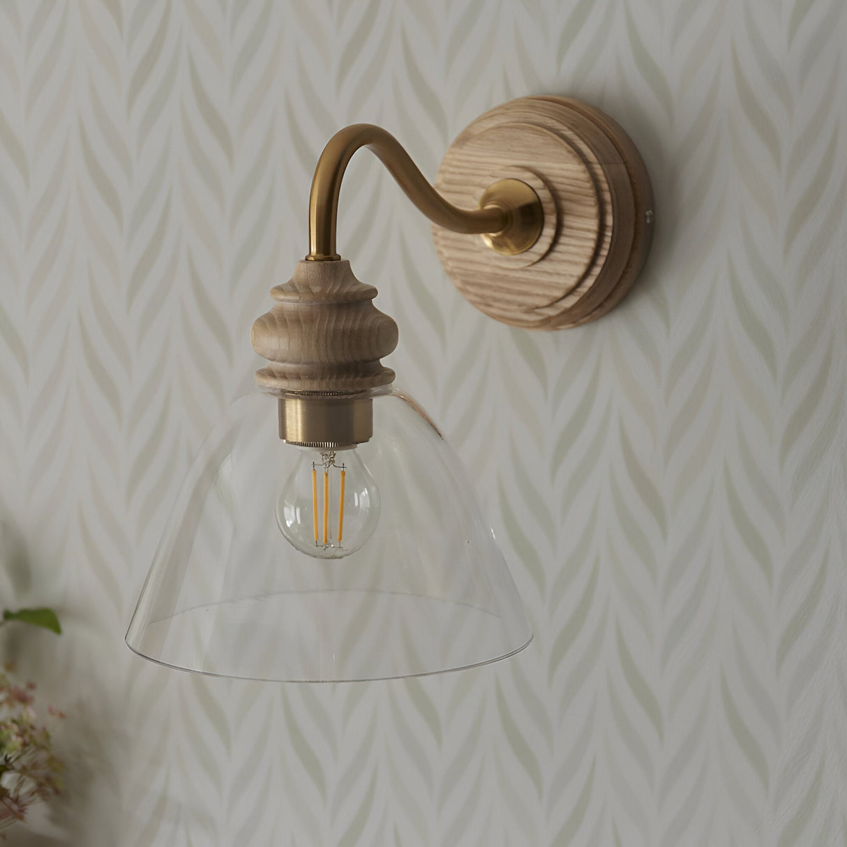 Endon Bevwick wall light in natural ash with aged brass curved arm and clear flared glass shade, mounted on patterned leaf-motif wallpaper.
