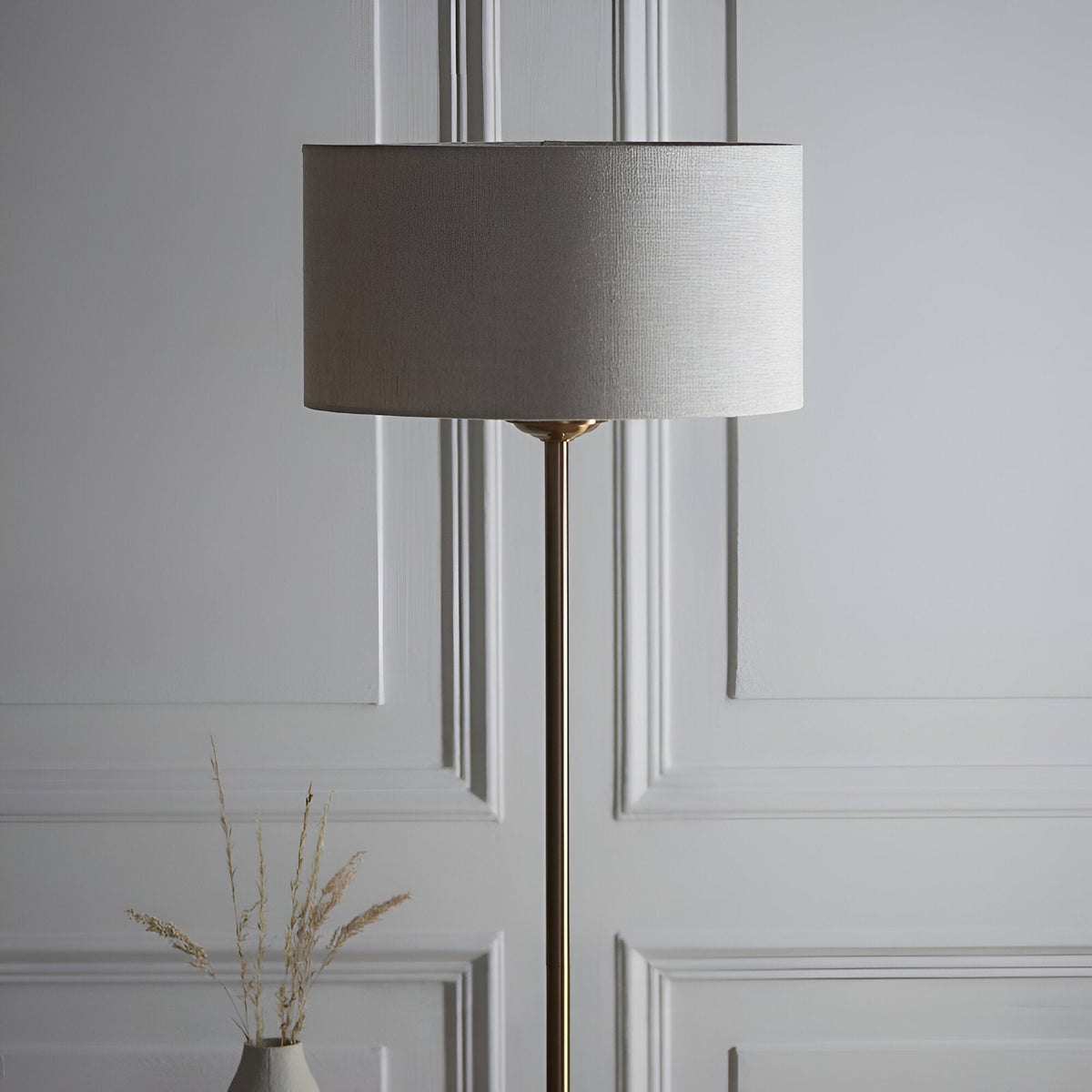 Brenley Floor Lamp in brass with a slim stem base and drum-shaped fabric shade, shown against a light grey panelled wall with a decorative vase nearby.