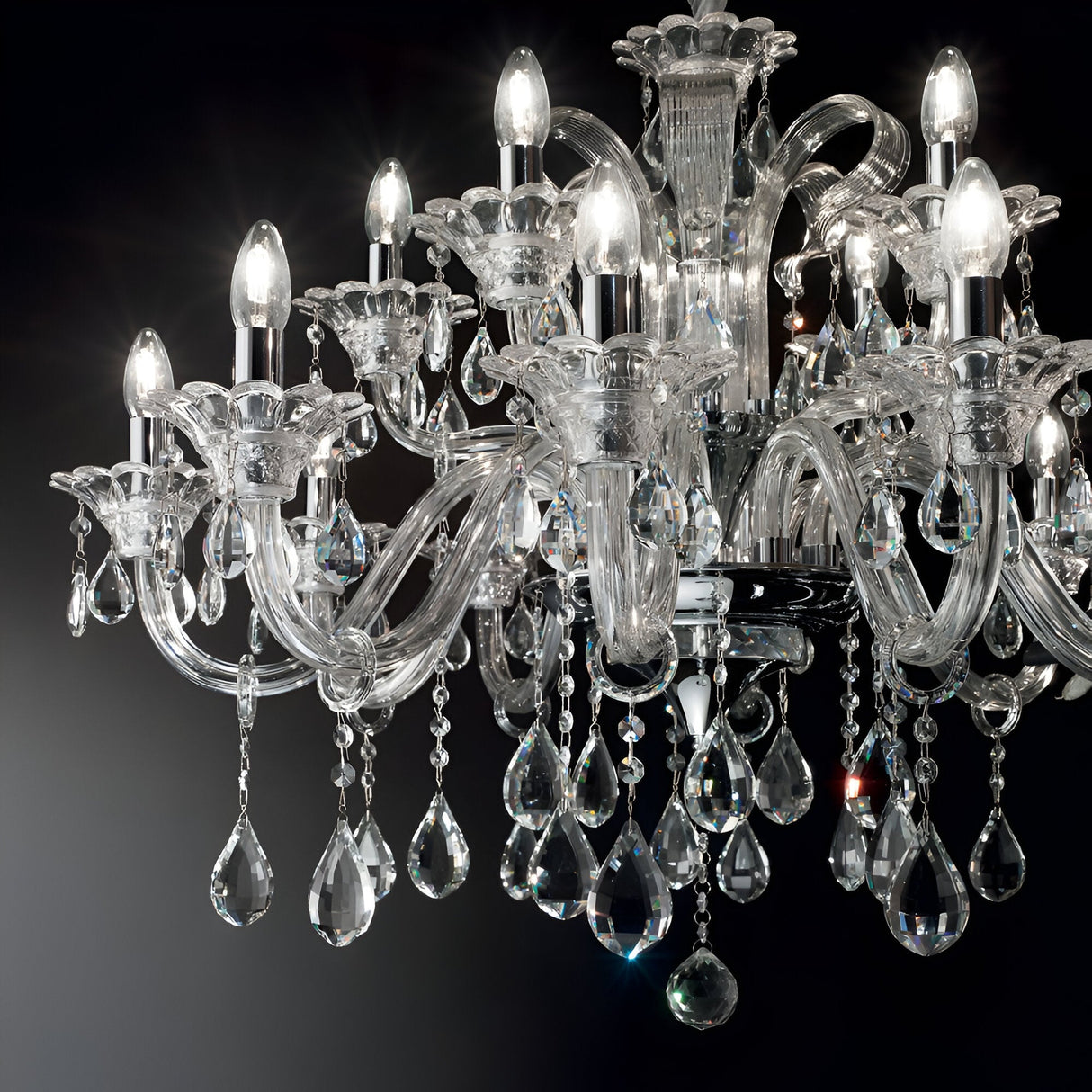 The Vivid Crystal 15 Light Chandelier - Chrome, featuring multiple arms and teardrop-shaped crystals, is lit against a dark background. The sparkling effect of the decorative lighting enhances the intricate glasswork and elaborate design.