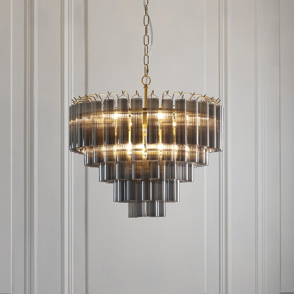 Endon Cranshaw 12 light tiered pendant with smokey grey cylindrical glass on a satin brass frame, suspended by chain against a pale panelled wall.
