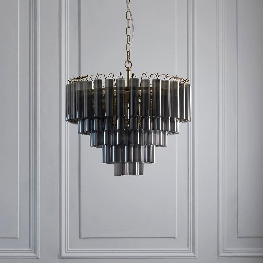 Endon Cranshaw 12 Light Tiered Pendant in satin brass with smokey grey cylindrical glass tubes, hung from a chain against a light grey panelled wall.