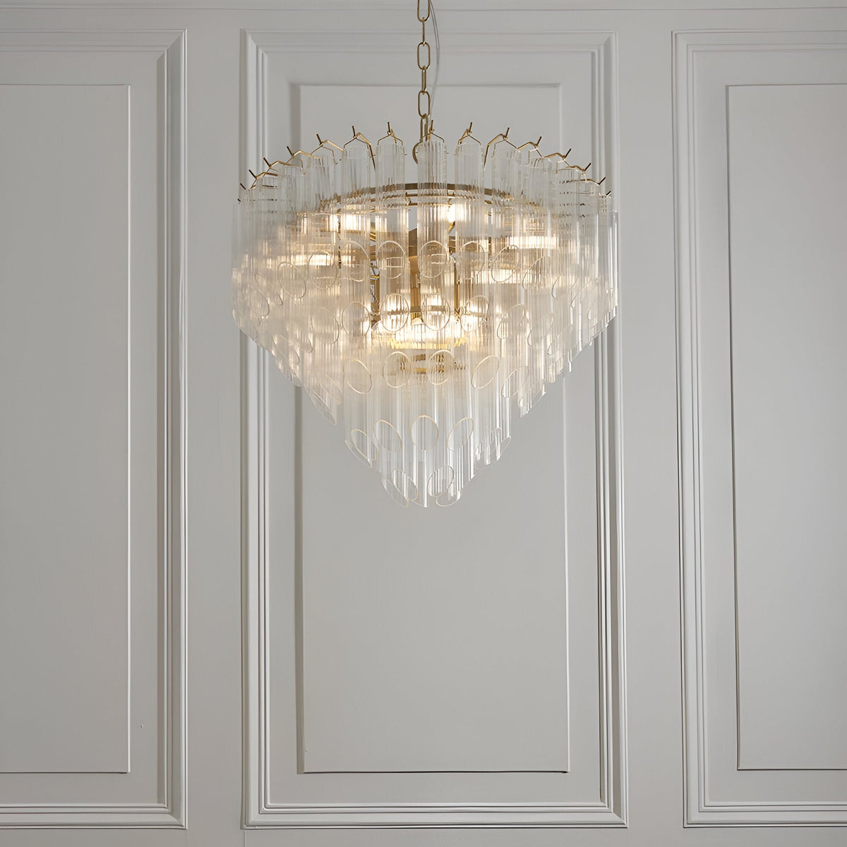 Cranshaw 12 Light Tiered Pendant in brass, with clear ribbed glass rods layered around the frame, hung from a chain against a panelled white wall.