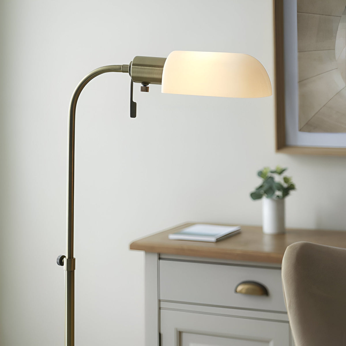 Endon Chartbourne floor lamp with antique brass curved stem and gloss opal glass shade, shown beside a wooden desk in a bedroom setting.