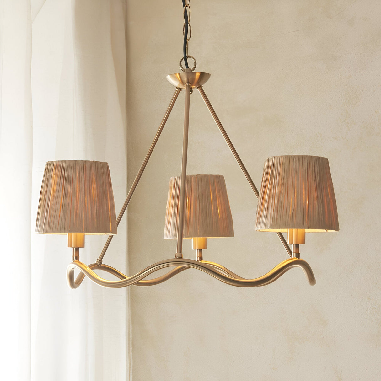 Danemont 3 Light Pendant in brass with a wave-shaped hoop and three hand-wrapped raffia shades, hung on a chain against a neutral wall.