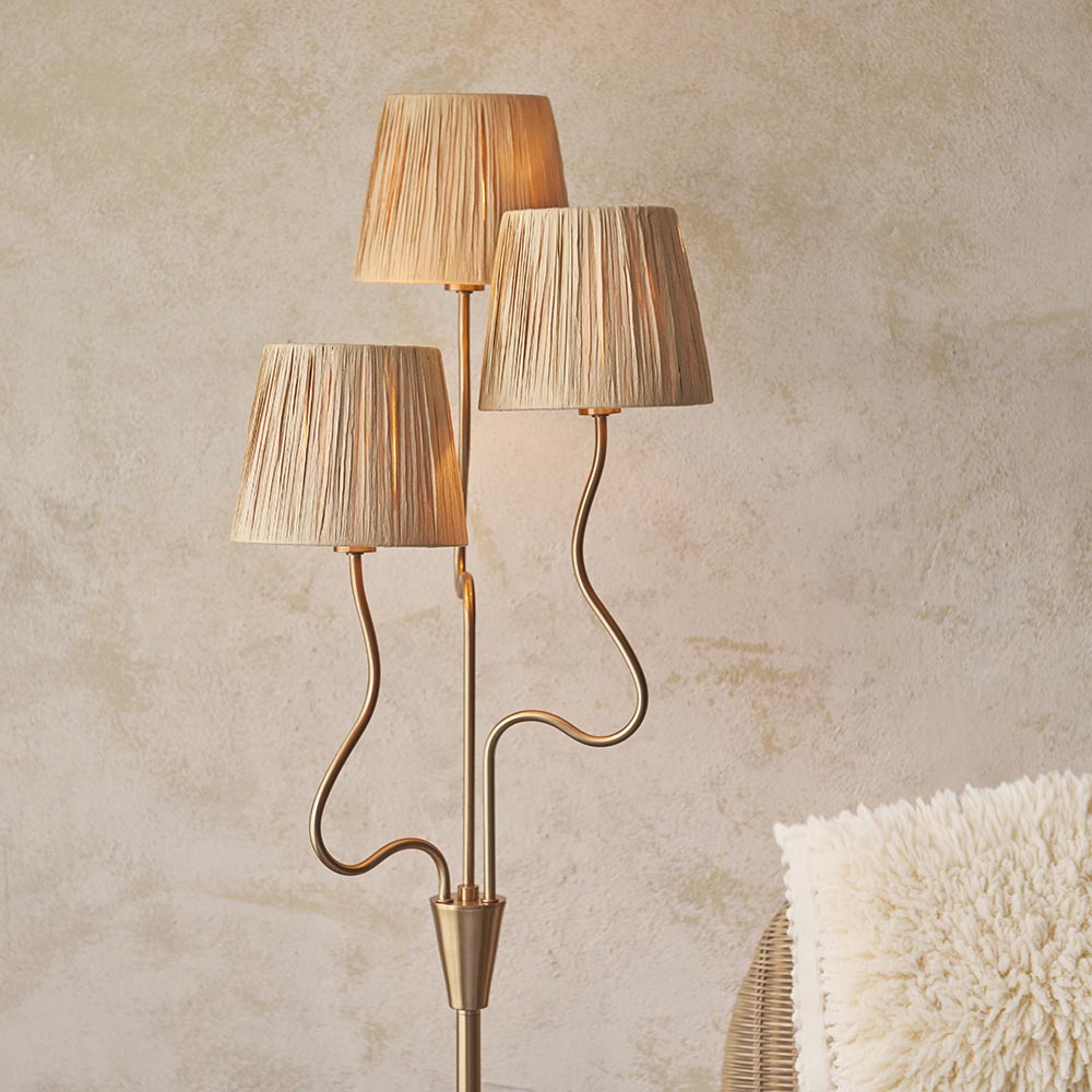 Danemont 3 Light Floor Lamp in aged brass with wriggle stem and three tapered raffia shades, shown in a room with a woven chair and cushion.