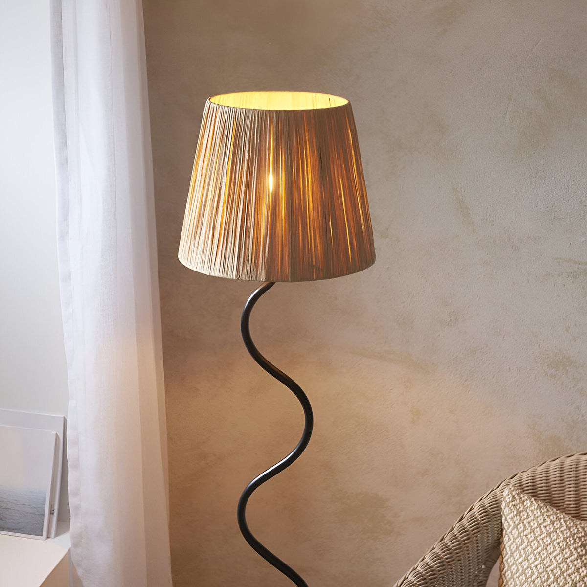 Endon Danemont floor lamp with matt black wriggle stem and tapered hand-wrapped raffia shade, illuminated in a bedroom corner beside a rattan chair.