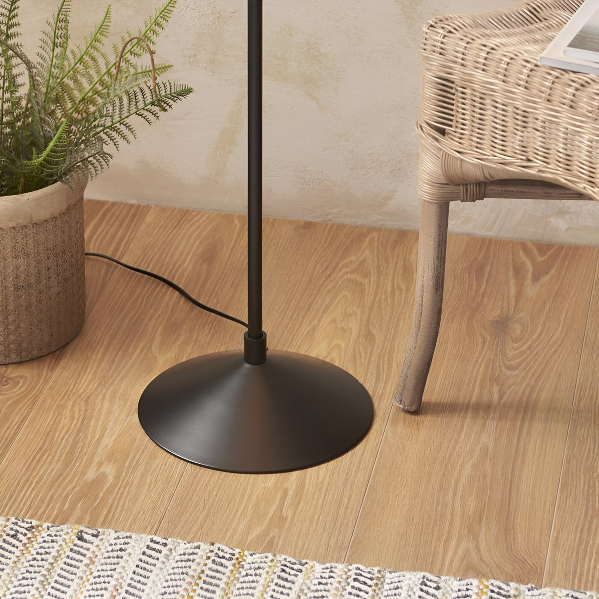 Close-up of the matt black round metal base and stem of the Endon Danemont floor lamp on wood flooring beside a rattan chair and potted fern.