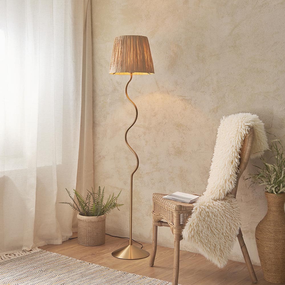 Endon Danemont floor lamp in aged brass with a wriggle-shaped stem and tapered raffia shade, styled beside a rattan chair and fern in a warm neutral room.
