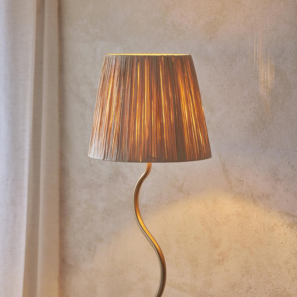 Endon Danemont floor lamp with a wriggle-shaped aged brass stem and tapered hand-wrapped raffia shade, lit beside a pale plaster wall.
