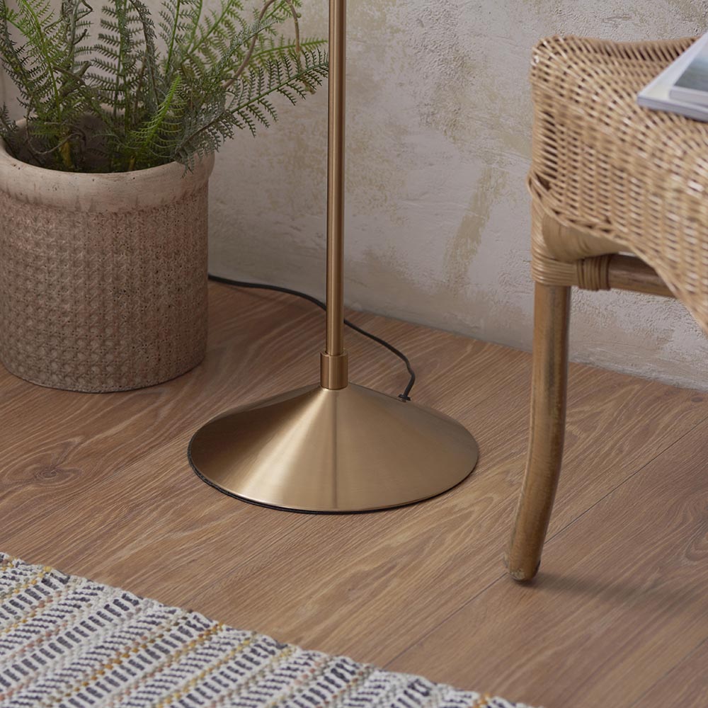 Close-up of the Endon Danemont floor lamp's aged brass tapered base and stem on wood flooring beside a rattan chair and potted fern.