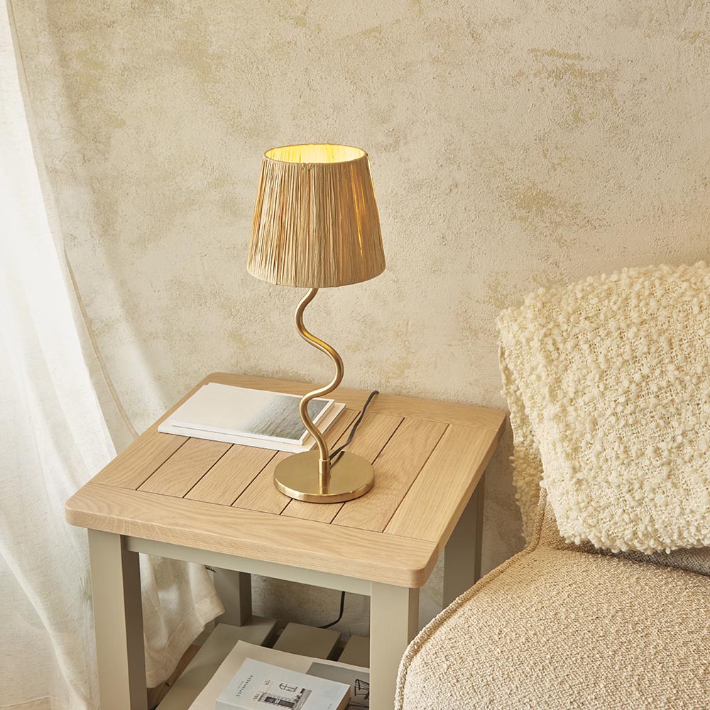 Endon Danemont table lamp with a curved brushed brass stem and tapered natural raffia shade, lit on a wooden side table beside a boucle chair.