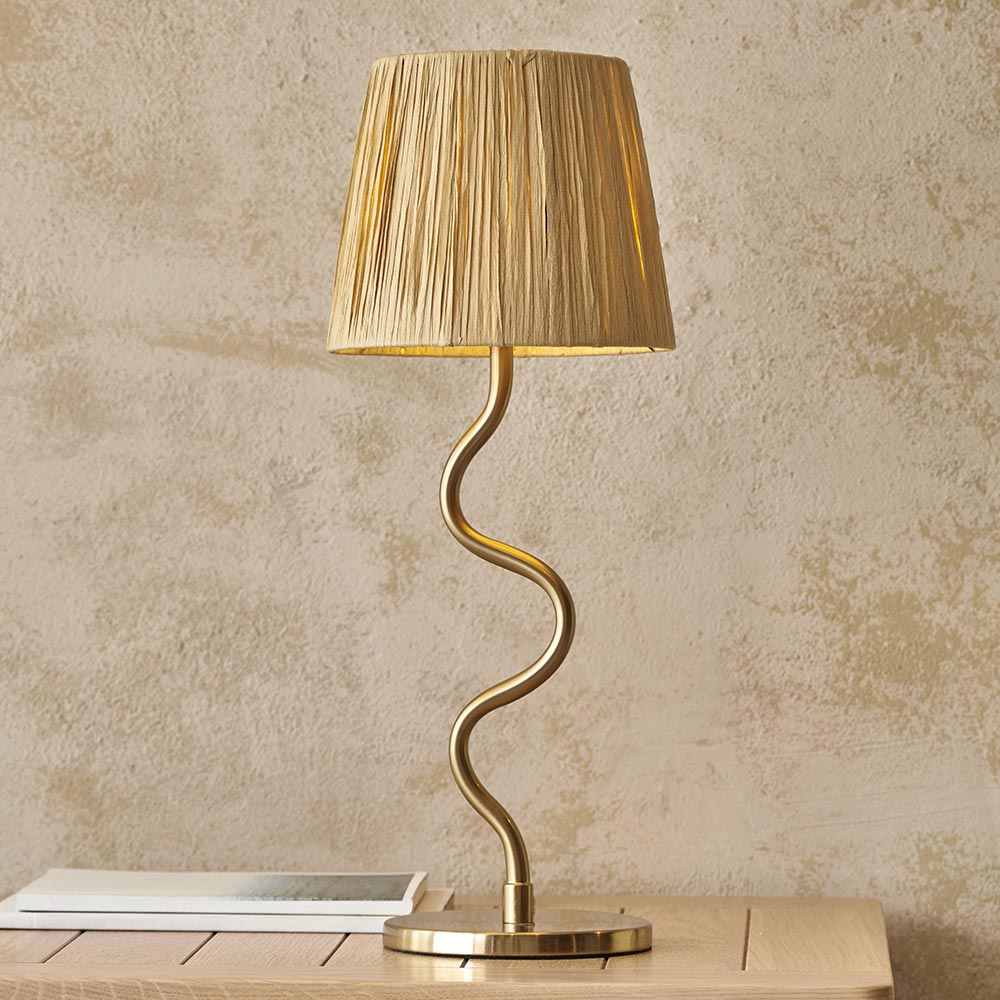Endon Danemont table lamp with a wavy brushed brass stem and tapered natural raffia shade, on a wooden surface against a textured beige wall.