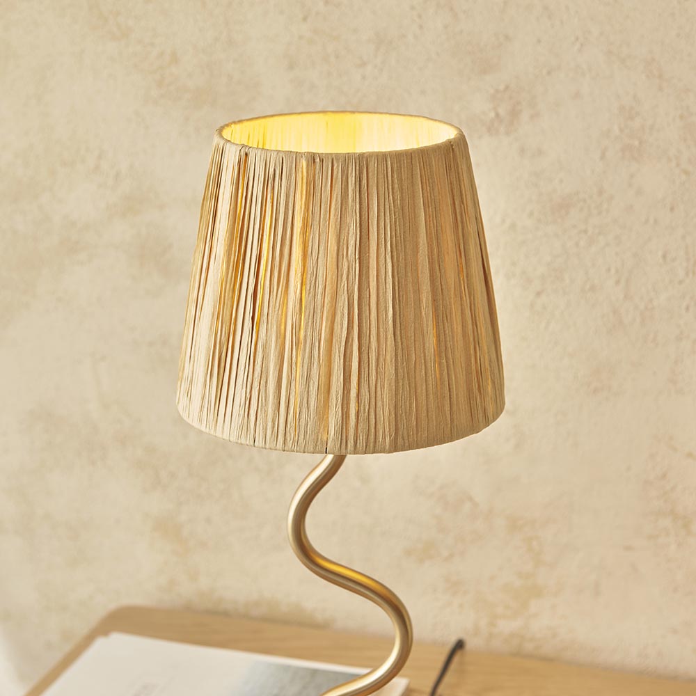 Endon Danemont table lamp lit on a wooden surface, with a tapered natural raffia shade on a wavy brushed brass stem against a neutral wall.