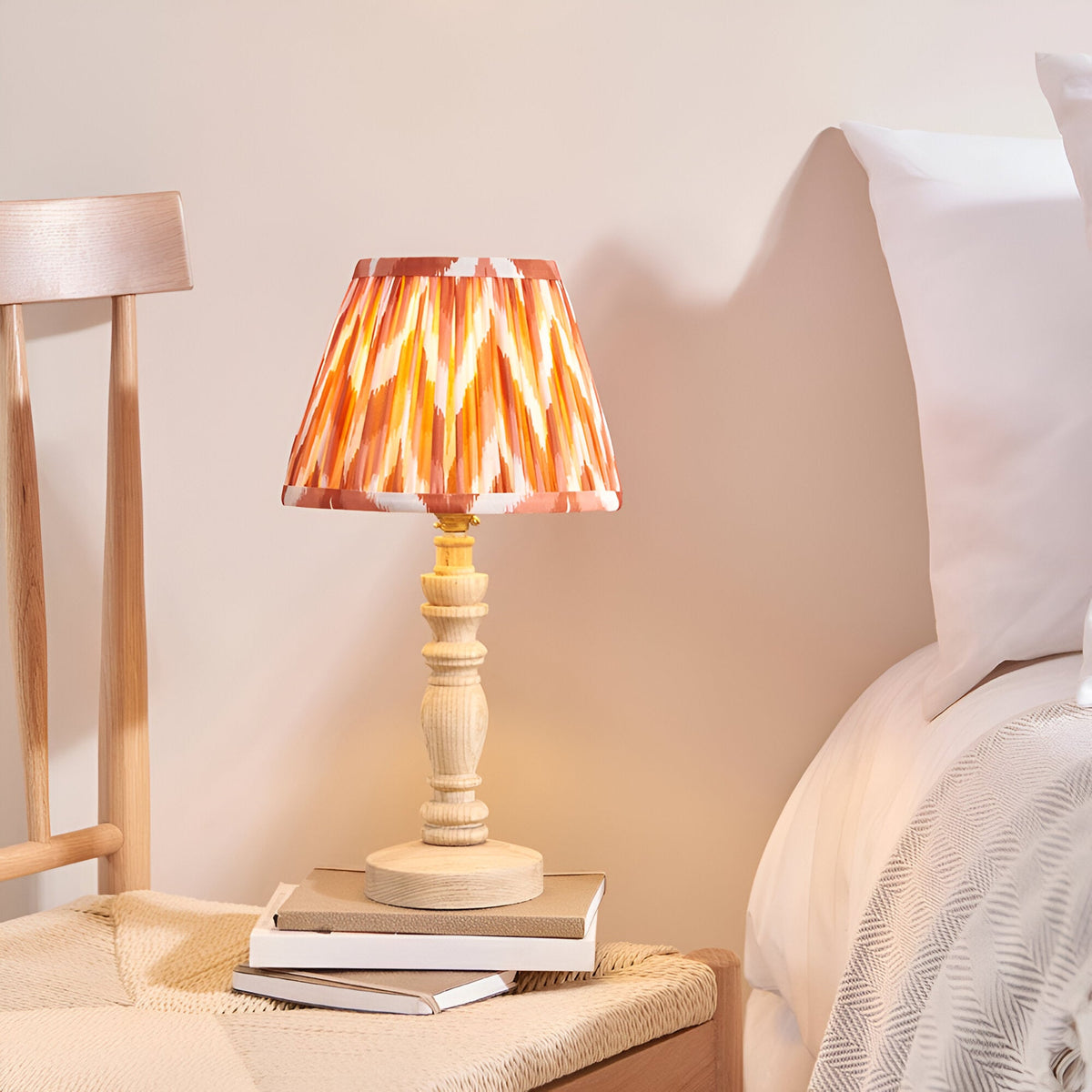 Endon Aldworth table lamp with turned natural wood spindle base and pleated zigzag shade in orange tones, on books beside a bed.