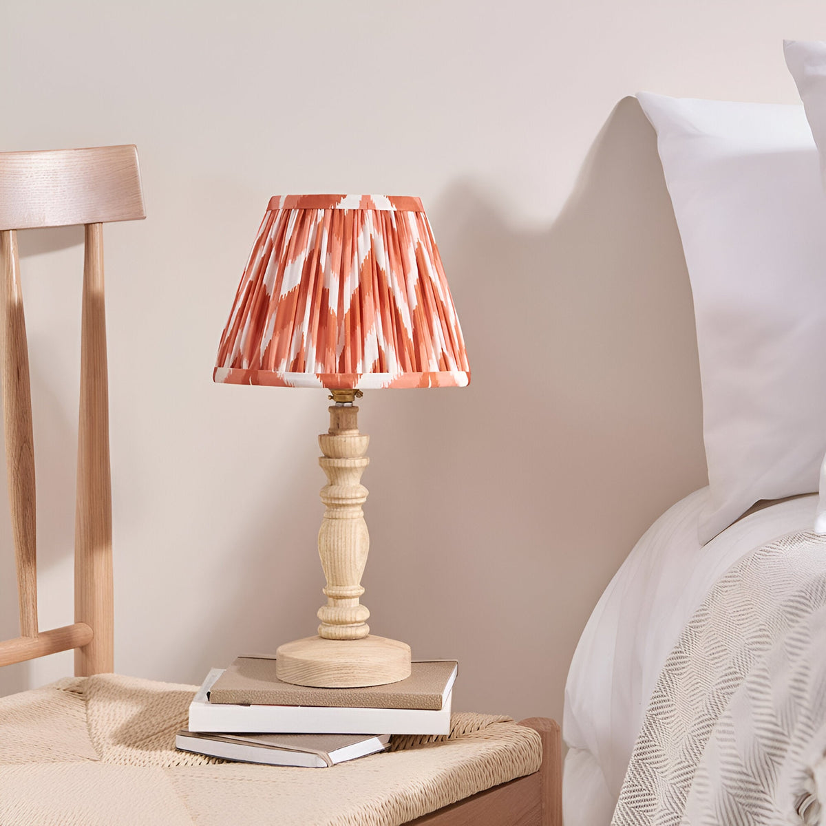 Endon Aldworth table lamp with turned natural wood base and pleated coral zigzag faux silk shade, on books beside a bed with white linens.