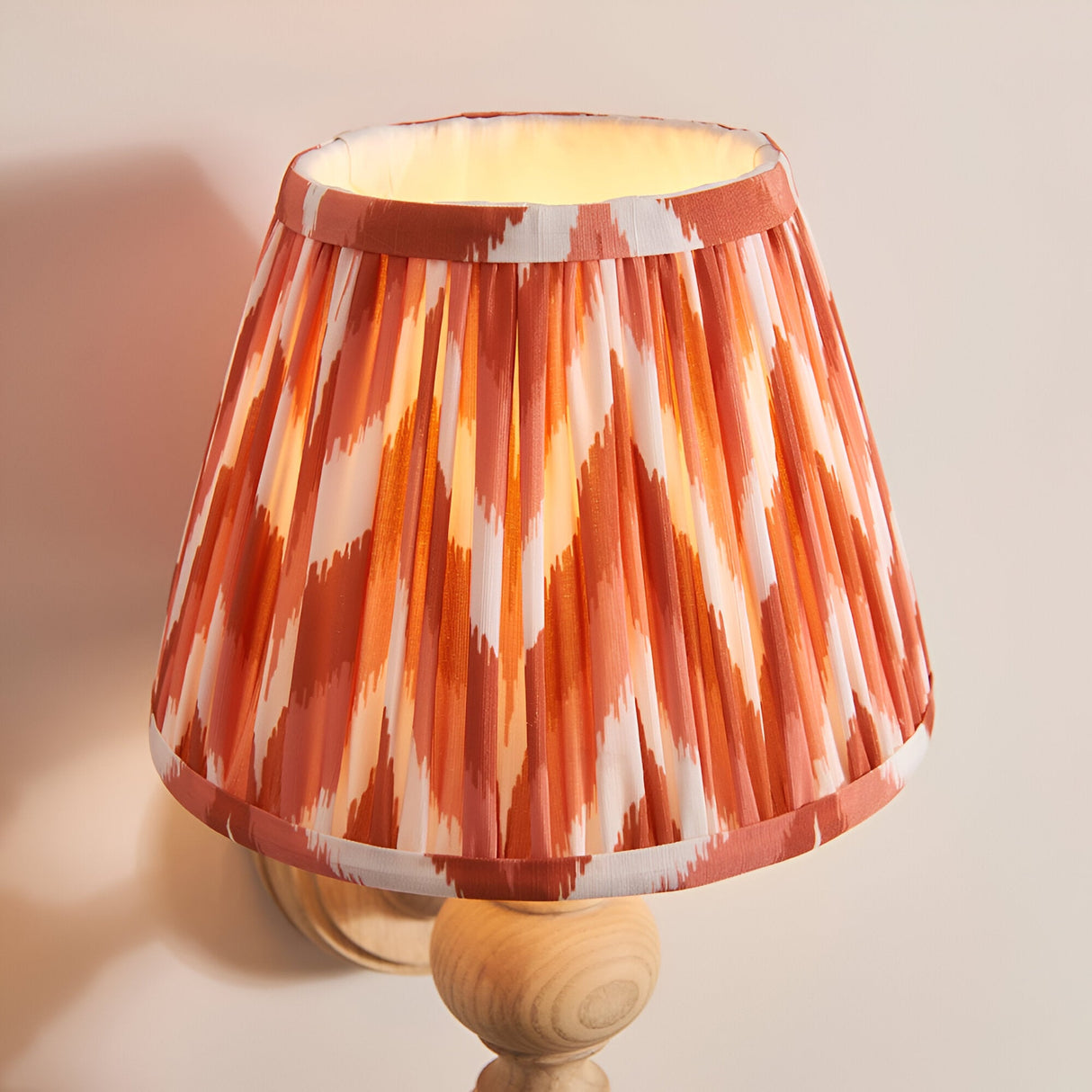 Aldworth Zigzag Wall Light in natural ash wood with turned spindle base and pleated apricot orange zigzag shade, glowing on a plain wall.