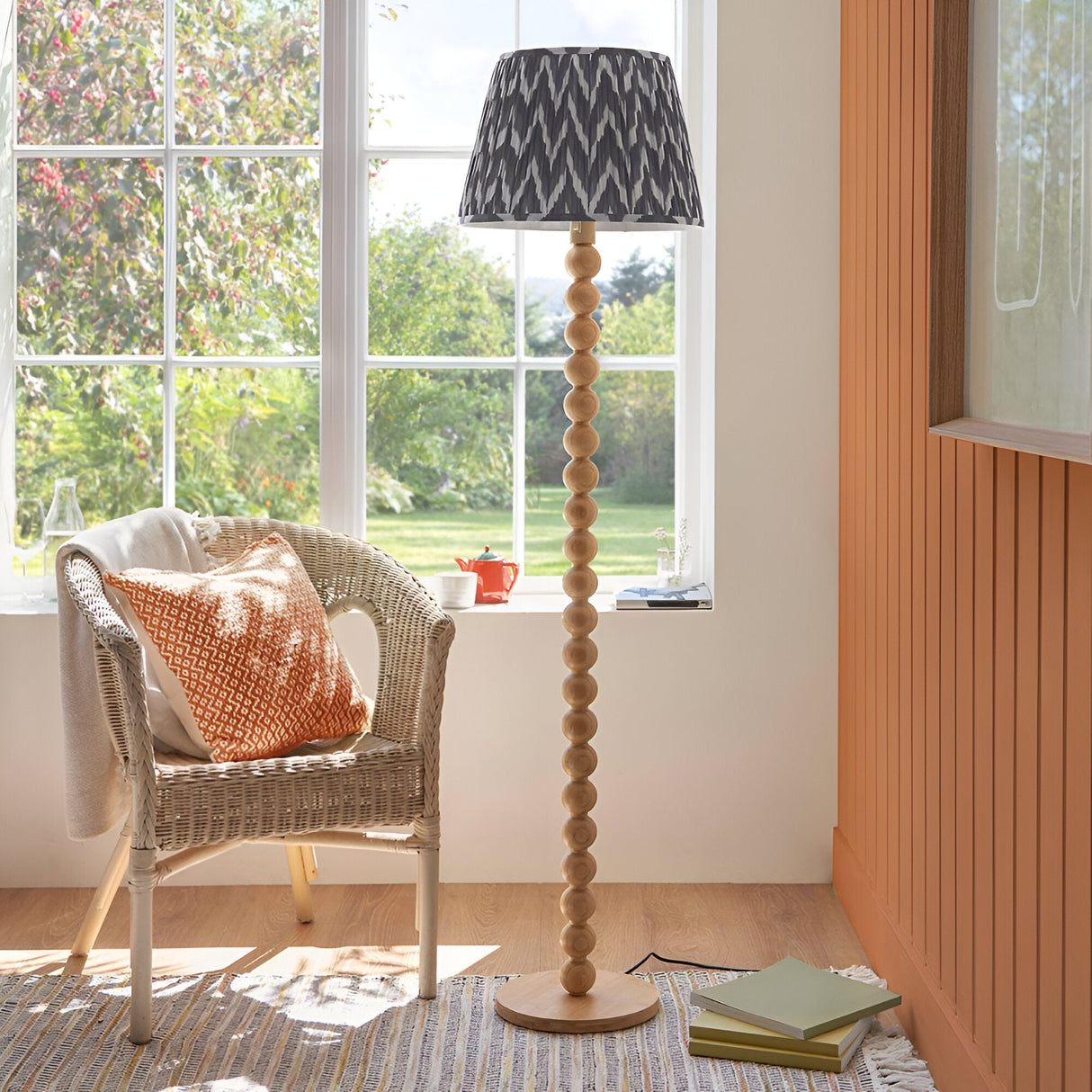 Ashwood Zigzag Floor Lamp in Grey with stacked wooden sphere base and pleated tapered shade, shown beside a wicker chair near a window.